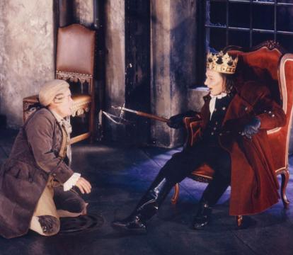 An actor in a blonde wig and brown coat kneeling in front of a king.