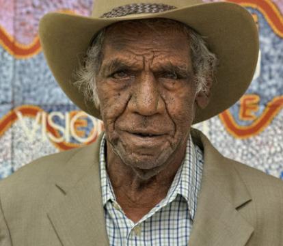 Michael Nelson Jagamara wearing a beige suit jacket over a blue and white checked shirt and wide brim hat, standing in front of an artwork in pastel colours.