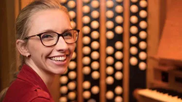 A smiling person wearing glasses sits at an organ, with music sheets.