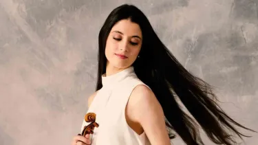 A woman with long, dark hair wearing a sleeveless white top holds a violin.
