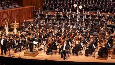 Simone Young stands with arms to the side in front of the Sydney Symphony Orchestra with a full choir insider the Concert Hall