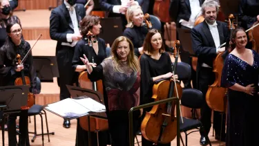 A woman in her 60s with long brown hair stands in front of an orchestra at the conductors desk, waving to the orchestra.