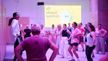 A dance cteacher stands with hands on hips to a room full of dance participants in pink lighting.
