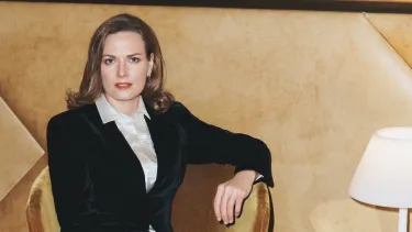 A woman in a ivory shirt and black blazer sitting in a velvet chair next to a side table with a lamp on it.