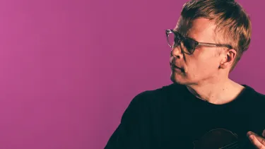 A man wearing a black t-shirt and glasses holding a violin against a magenta coloured background.