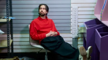 A white man sits in a chair with his feet up wearing a red sweater.