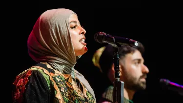 A woman in a colorful hijab speaks passionately into a microphone on stage, accompanied by a man in traditional attire.