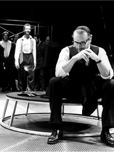 Actors on stage, a man at the front in glasses and a hat.