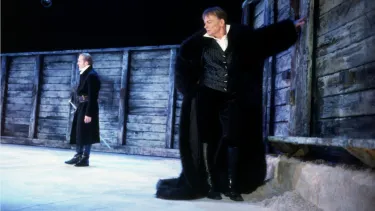 Two actors in long black coats stood next to a wall on stage.