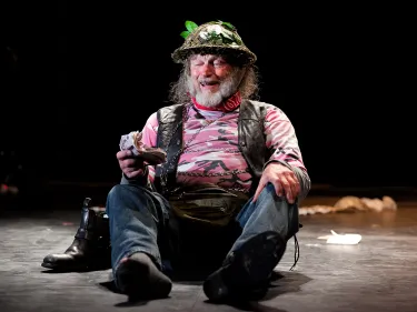 An actor in a pink camoflauge top and leafy helmet sat on stage.