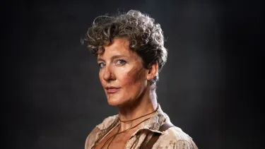 Calamity Jane: Virginia Gay with short curly hair and dirt on their face wearing a beige shirt with open collar, looking at the camera against a dark background.