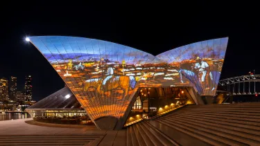 The Sydney Opera House sails at night, illuminated with an Australian outback landscape in orange and browns with a blue sky.