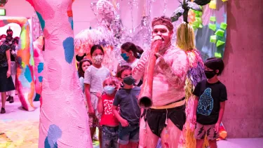 A First Nations man on a didgeridoo in the centre for creativity leads a line of children in a pink room.