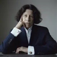 A mature white woman staring at the camera with a slight frown. She is wearing a navy suit, her hair is curly and ends at her jaw and she is leaning on her right hand as if in thought.