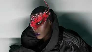 A black woman wearing a striking red butterfly mask adorned with white dots, set against a dark, textured outfit with a high collar.