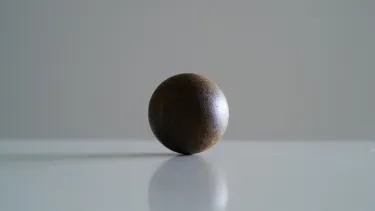 A small brown metal ball sits on a white surface. The background is blured.