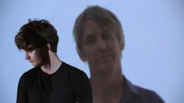 Joe Keery looks to the left as Stephen Malkmus is projected over his shoulder.