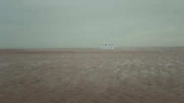 In a barren desert with a grey cloudy sky, a group of people stand in the distance in all white clothing.