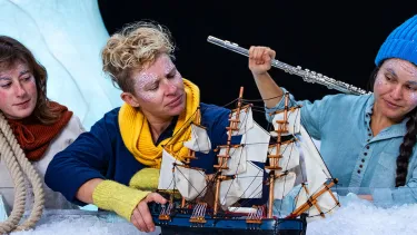 Three people with frosty face paint engage with a model ship on artificial snow. One holds a rope and another a flute, set against icy backdrops, creating a whimsical arctic scene.