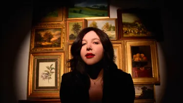 A woman with brown hair looks at the camera standing in front of a wall of gold picture frames.