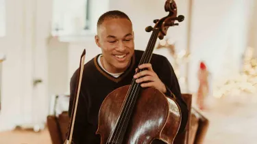 Sheku Kanneh-Mason holds a Cello in front of a piano.