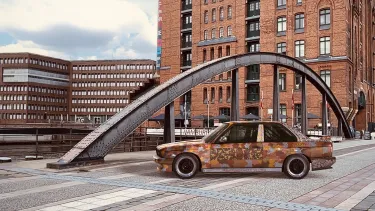 A BMW sedan covered in patterns of ochre, yellow, brown and blue parked on the pavement in front of high-rise buildings.