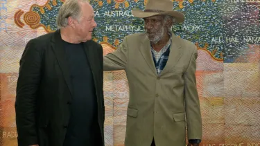 Two men looking at each other, one with his arm on the other's back, standing in front of a pastel coloured artwork.