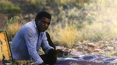 Michael Nelson Jagamara sitting on the ground, surrounded by bushland, leaning over a painting, laid out in front of him.