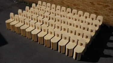 A group of 60 cream chairs with a square cut out in the back sit angled to the left in the sun infront of a brown bricked wall.