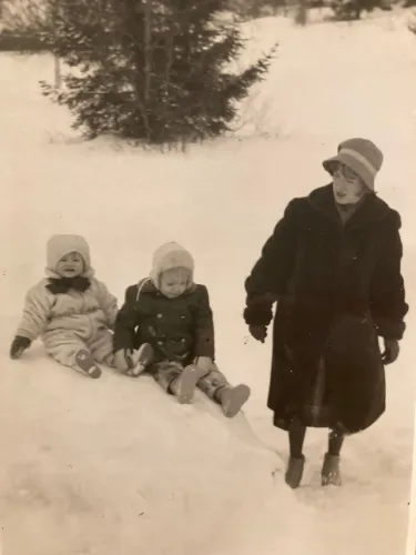 Two children sliding in the snow behind a young woman in a coat.