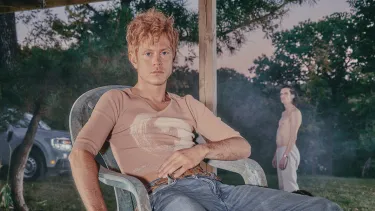 A white man wearing a beige top and baggy jeans louges in a plastic chair.