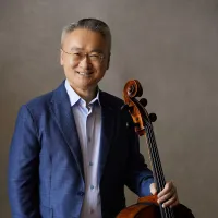 A Chinese man with short greying hair wears a blue suit and stands holding a cello.