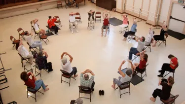 A group of seated people in a studio, gathered in a circle with their hands above their heads.