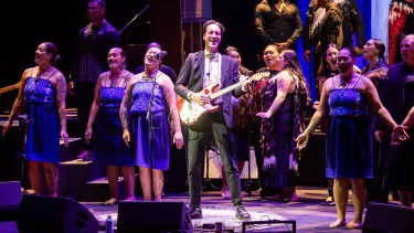 Marlon Williams on stage holding a guitar in a grey suit with several backing singers.