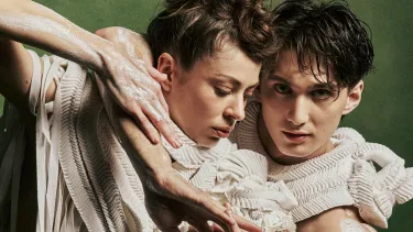 Two dancers in off-white textured costume with arms interlocked and white clay on their bare arms, against a dark green background.