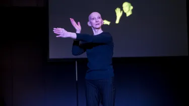 The artist makes hand symbols to the side with the same hand symbols projected to a screen behind them.