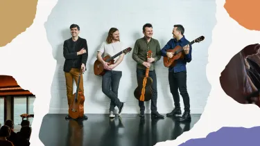 Four men holding guitars stand casually against a white brick wall. They are smiling and interacting.