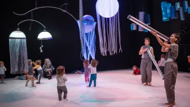 A group of young children plays under jellyfish-like lights on a stage, with adults engaging them using tubular instruments.