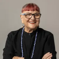 A white woman in her 60s wears glasses and a black shirt. She has dyed red hair with a short fringe and smiles at the camera.
