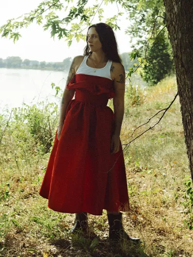 Woman in red dress standing by river, gazing into distance.