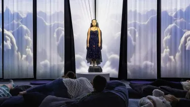 A young white woman with dark brown hair stands in front of a fabric background, where projections of clouds are being cast. An audience of meditation children and adults lie on pillows in front of her.