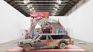 A car adorned with vibrant paint splashes and various decorative items, on display in a white gallery.